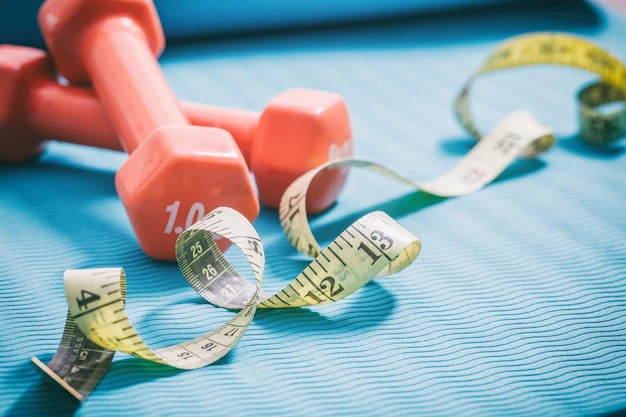 A pair of dumbbells placed on the floor with an inch tape beside them. The dumbbells are made of metal and have a textured grip for better handling. The inch tape, with measurements marked on it, is rolled out next to the dumbbells.