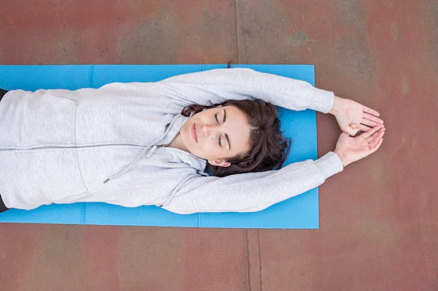 A girl lying on an exercise mat, positioned in a relaxed and peaceful manner. Her body is stretched out on the mat, with her arms raised straight above her head and her hands gently clasped together.