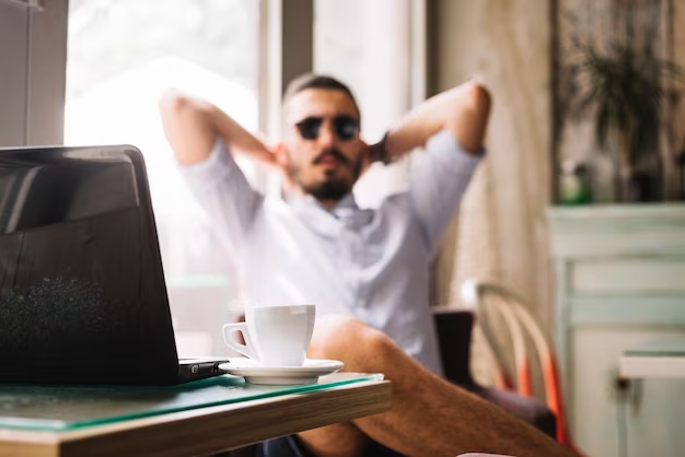 A handsome man sitting comfortably with his back supported on a chair, in front of a laptop. A cup of tea is placed on the table within his reach to make him feel positive.